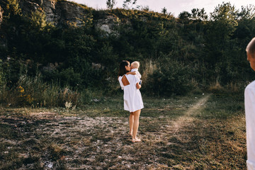 Walk beautiful young family in white clothes with a young son blond in mountainous areas with tall grass at sunset. Mother keeps son in her arms, hugging. family - this is happiness