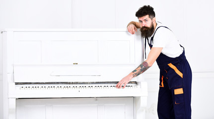 Man with beard and mustache, worker in overalls lean on piano, white background. Loader moves piano...