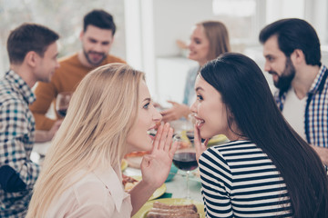 Two attractive, stylish, best girls holding palm near face whispering girlish gossips to each other about their friends who sitting at the table enjoying dinner, supper