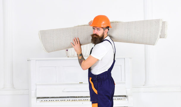 Relocating concept. Man with beard, worker in overalls and helmet carries rolled carpet, white background. Loader wrapped carpet into roll. Courier delivers furniture in case of move out, relocation.