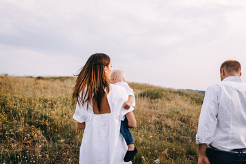 Walk beautiful young family in white clothes with a young son blond in mountainous areas with tall grass at sunset. Mother keeps son in his arms, hugging. family - this is happiness