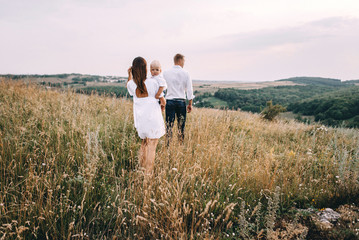 Walk beautiful young family in white clothes with a young son blond in mountainous areas with tall grass at sunset. Mother keeps son in his arms, hugging. family - this is happiness