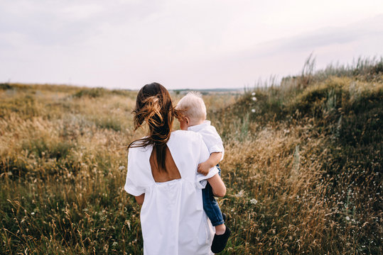 Walk beautiful young family in white clothes with a young son blond in mountainous areas with tall grass at sunset. Mother keeps son in his arms, hugging. family - this is happiness