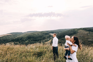 Walk beautiful young family in white clothes with a young son blond in mountainous areas with tall grass at sunset. Mother keeps son in his arms, hugging. family - this is happiness