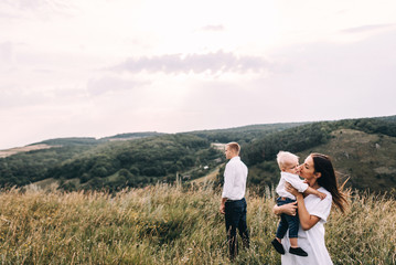 Naklejka premium Walk beautiful young family in white clothes with a young son blond in mountainous areas with tall grass at sunset. Mother keeps son in his arms, hugging. family - this is happiness