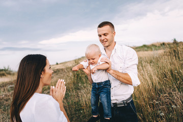 Fototapeta premium Walk of beautiful young family in white clothes with a young son blond in mountainous areas with tall grass at sunset. Dads carries his son on his shoulders. family - this is happiness