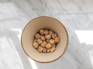 Top view of a coffee-colored ceramic cup containing a handful of toasted and peeled hazelnuts. The cup is placed on a marble shelf illuminated by the sun's rays