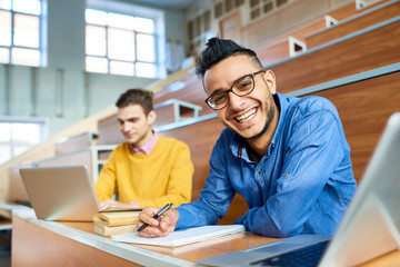 Fototapeta premium Portrait of two students sitting at desks in modern auditorium at college and preparing for class, focus on young Middle-Eastern man looking at camera and smiling, copy space