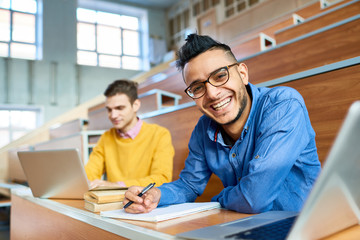 Portrait of two students sitting at desks in modern auditorium at college and preparing for class,...