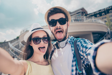 Foolish crazy comic funny couple in caps having journey trip meeting outdoors shooting self portrait on front camera showing tongue out, beautiful and handsome bloggers having video-call with friends