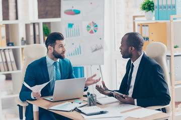 Stylish, attractive, handsome employees in classy suit with tie sitting in work place, using tablet, laptop, gesturing with hands, talking about new project, having papers, documents on the table