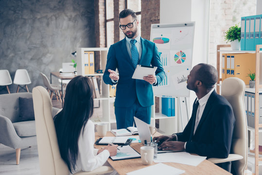 Handsome, stylish, attractive, smart coach in classic suit with tie holding tablet gesturing with hand telling to his colleagues about startup, making consultation, standing in work station, place