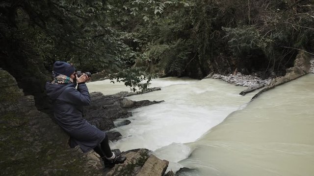 a young woman in warm clothes holds a camera in her hand, a photographer takes pictures of a beautiful waterfall, water falls from above, the lady likes a natural phenomenon