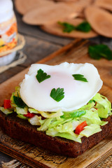 Yummy poached egg sandwich. Fast poached egg on rye bread slice with fresh vegetable mix and parsley. Healthy breakfast food to help lose weight. Rustic wooden background. Closeup