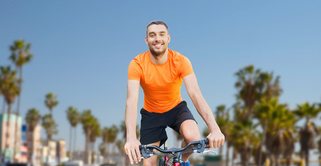 Fototapeta premium fitness, sport and healthy lifestyle concept - happy young man riding bicycle over venice beach background in california