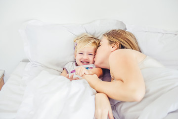 Obraz na płótnie Canvas Happy and loving mother having fun and kissing her daughter on the bed. Family morning concept. Spending free time together at home. Soft selective focus. Space for text.