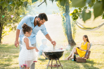 Side view of a cute girl watching her father preparing meat on a round charcoal barbecue grill outdoors during family picnic in a summer day - obrazy, fototapety, plakaty