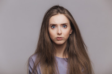 Studio shot of pleasant looking blue eyed female model with dark long hair, has puzzled expression as hears shocking news from interlocutor, isolated over grey background. Facial expressions concept