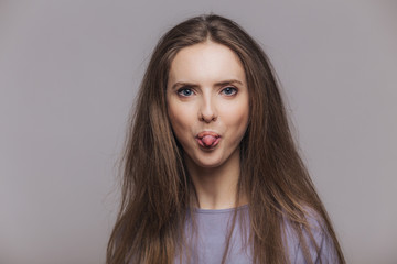 Funny brunette female with atractive look, shows tongue, makes grimace and has fun indoor, looks with blue eyes at camera, has dark straight hair, isolated over grey background. Positivity concept