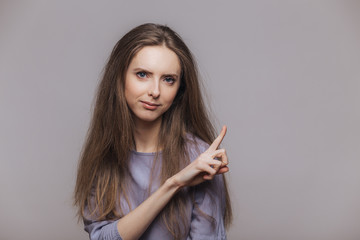 Fototapeta premium Cute young female model with brunette long hair, points at blank copy space of grey wall, advertizes something with serious expression, attracts your attention. Advertisment and people concept