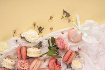 Pink and white macaroons cakes with big and small flower buds are decoratively laid on a white fabric on a yellow background