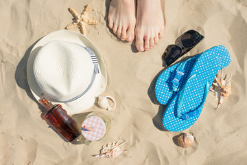 Hat, sunscreen, slippers, legs and seashells on sand, top view