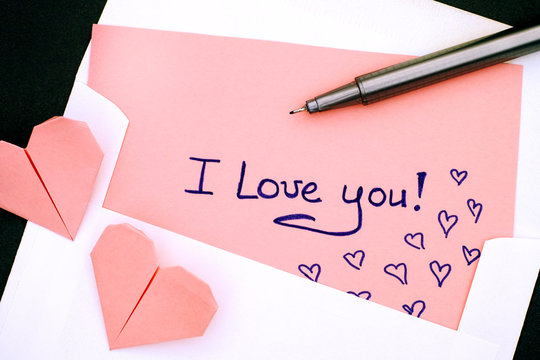 Letter with text I Love You! in envelope with two pink origami hearts and pen.