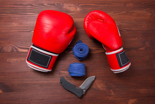 Red boxing gloves, bandage and folding knife on wooden brown background. Top view.