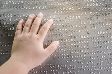 The blind kid's hand and fingers touching the Braille letters on the metal plate to understand an...