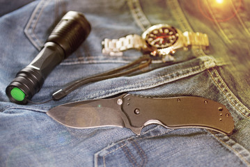 Folding knife, searchlight and men's watch on the jeans backgound