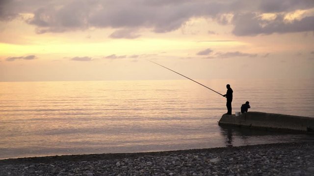 Silhouettes of father and his son fishing together at a seashore early in the morning. Locked down real time establishing shot