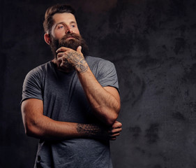 Portrait of a hipster with full beard and stylish haircut, dressed in a gray t-shirt, stands with a thinking look in a studio on a dark background.