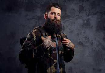 A handsome bearded traveler, dressed in a trendy camouflaged jacket, with a tattoed arms, standing in a studio, isolated on a dark background.