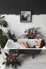 Sexy and beautiful young model woman with red hair and bright makeup, in black stylish bodysuits, mesh pantyhose and black boots, lies in bath with closed eyes