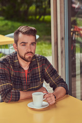 A handsome fashionable male with stylish haircut and beard, wearing fleece shirt, drinking coffee at the cafeteria.