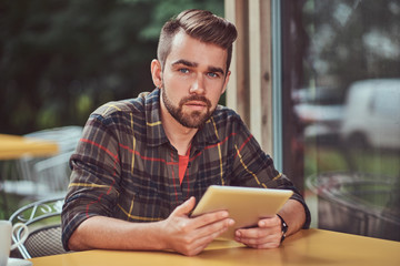 A handsome fashionable male freelancer with stylish haircut and beard, wearing fleece shirt, working on a tablet computer inside a cafe.