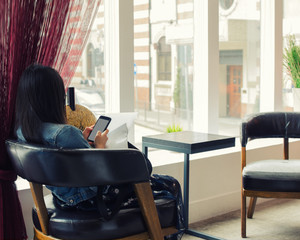 Young woman with dark hair sits at table near the window in hotel lobby and uses mobile phone.