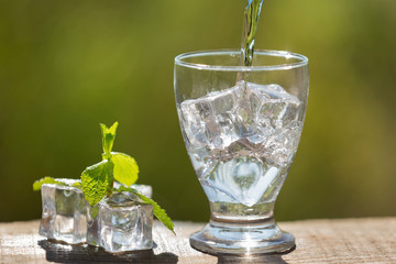 a glass with ice cubes pour soda water from a bottle, nearby lie ice cubes and mint, in the open space, sunlight and bokeh