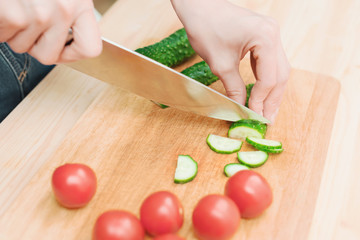 close-up of female hands cut into fresh cut cucumbers on a wooden cutting board next to pink tomatoes. The concept of homemade vegetarian cuisine and healthy eating and lifestyle