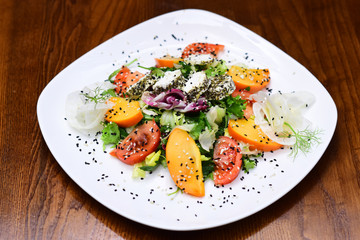Salad served in white plate, dark wooden background. Restaurant dish concept. Dish in restaurant salad with tomatoes, arugula, persimmon, onions, lettuce, radicchio, feta cheese with spices, sesame.