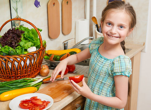 Child girl cooking in home kitchen, chopping tomatoes. Basket of vegetables and fresh fruits in kitchen interior. Healthy food concept