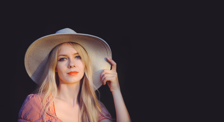 Beautiful young woman in pink dotted summer dress and sun hat points to copy space, looking at camera. Three quarters of a studio length shot on a dark natural background
