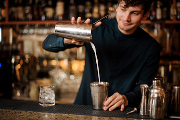 Fototapeta na wymiar Smiling brunett bartender pouring an alcoholic cocktail into a steel shaker