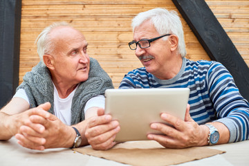 Portrait of two cheerful senior men using digital tablet sitting at table at outdoor terrace and chatting, copy space