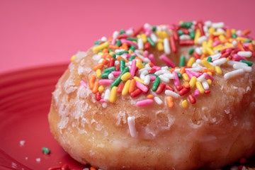 Close up of glazed doughnut with colorful sprinkles on a red plate with a pink background.