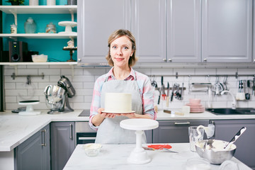 Portrait of professional female confectioner holding appetizing frosted cake in kitchen and smiling at camera