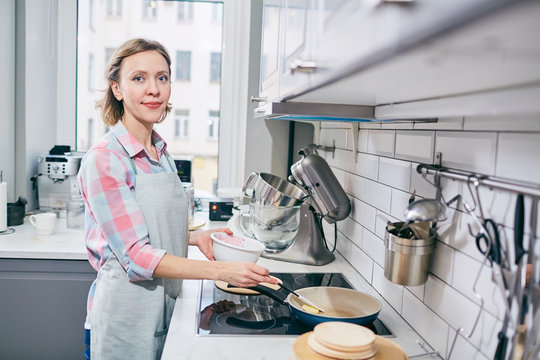 Portrait of attractive Caucasian woman standing by electric stove with frying pan in modern kitchen and smiling at camera