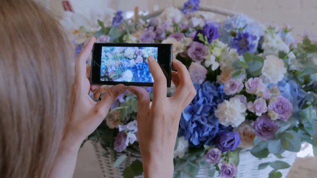 Professional floral artist, florist using smartphone and taking photo of her large floral basket with flowers at workshop, flower shop. Photography, technology and social media concept