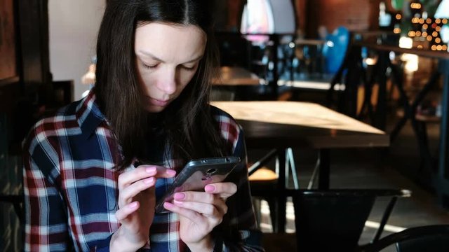 Upset woman sends a message or use internet in the phone sitting in a cafe. Dressed in a plaid shirt.