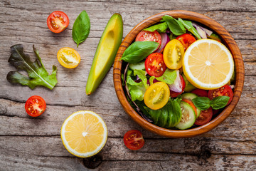 The bowl of healthy vegan salad . Various vegetables avocado, tomato, cucumber, red cabbage, basil and onion on shabby wooden background .